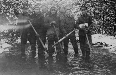 p of men standing in a river, dressed in winter clothing, holding axes.