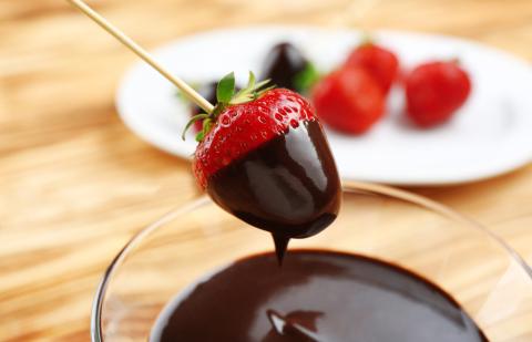 Image of a strawberry being dipped in chcolate fondue. There is a plate of strawberries in the backgound on a table.