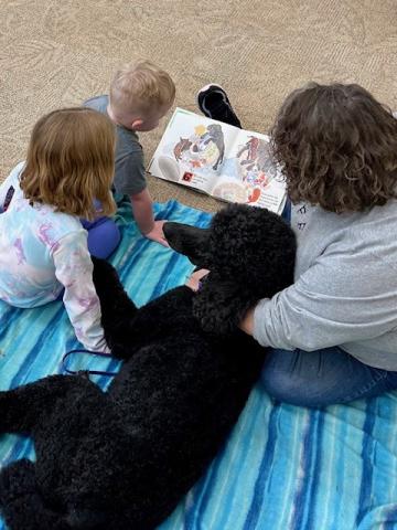  A mom, her daughter, and her son are sitting on a blanket on the floor with a black dog, reading a book together.