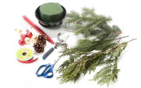 An image of evergreen tree branches laying on a counter with scissors, ribbons, pine cones, Christmas ornaments, and other wreath-making supplies.