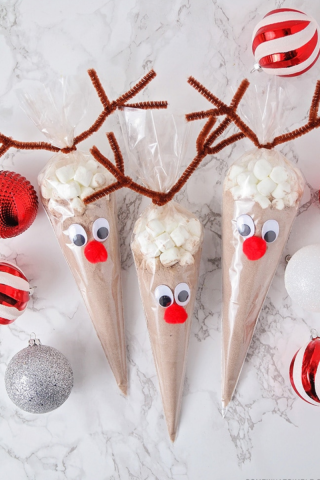 Image of three frosting bags filled with hot chocolate mix and mini marshamallows. The bags are made to look like reindeer with ppipe cleaner antlers, googly eyes, and round red noses. These are laying on a countertop surrounded by red and silver Christmas ornaments.