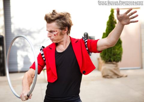 Image of a man with blond hair, and a hand in the air, twirling a large metal ring. He is wearing a black shirt and pants, with a red jacket.