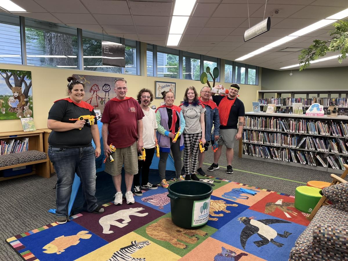 A group of adults with NERF guns in the library, standing in front of book shelves.