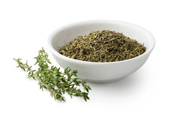 Image of a white bowl filled with thyme leaves and a full stem of thyme laying next to it.