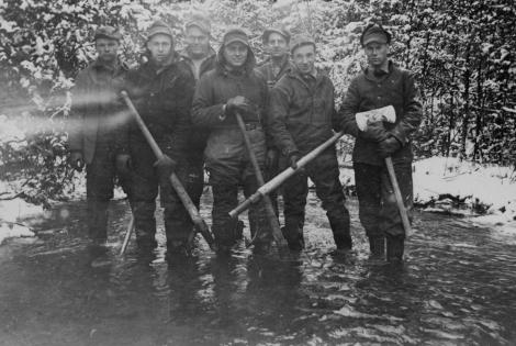 p of men standing in a river, dressed in winter clothing, holding axes.